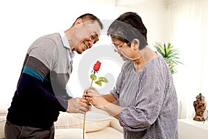 Romantic elderly man gives red rose flower to his wife in living room, happy smiling Asian senior mature retired couple celebrate