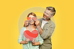 Romantic european middle aged man covering wife eyes with red heart shaped cards and smiling, yellow background