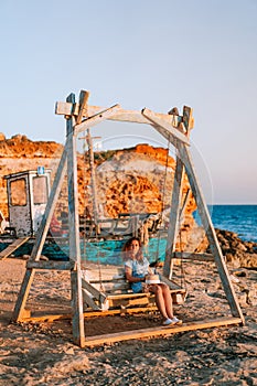 Romantic evening at sunset by the sea, a young woman in a dress with a bouquet of flowers is sitting on a wooden swing holding her