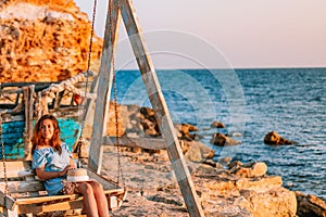 Romantic evening at sunset by the sea, a young woman in a dress with a bouquet of flowers is sitting on a wooden swing holding her