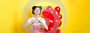 Romantic girl close eyes and pucker lips for kiss, showing I love you heart gesture, standing near cute red balloons