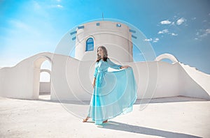 Romantic girl with curly hair in blue chiffon dress posing by traditional greek white houses. Beautiful model enjoying Mykonos