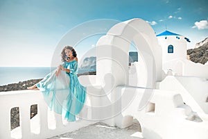 Romantic girl with curly hair in blue chiffon dress sitting on white balcony by arch over blue sky. Beautiful bride model enjoying