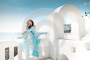 Romantic girl with curly hair in blue chiffon dress sitting on white balcony by arch over blue sky. Beautiful bride model enjoying