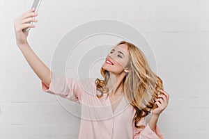 Romantic girl with shy smile making selfie while playing with blonde hair. Indoor portrait of charming young woman in