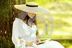 Romantic Girl in White Dress and Straw Sun Hat reading Book sitting under Tree. Young Woman studying in Spring Park. Old Fashioned
