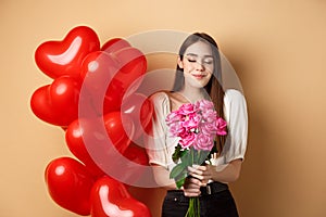 Romantic girlfriend smelling roses, receive flowers from lover on Valentines day, standing near red hearts balloons and