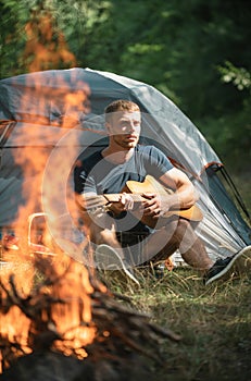 Romantic guy camping outdoors and sitting near tent. Handsome man playing guitar in forest with bonfire. Country music