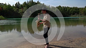 Romantic jazzman is playing music by saxophone on shore of calm lake in forest, summer day