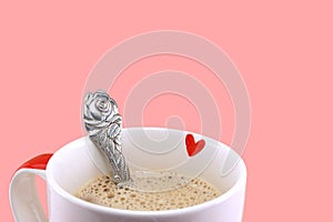 small red heart inside edge of white ceramic coffee cup and rose flower shaped metal silver spoon isolated on pink background