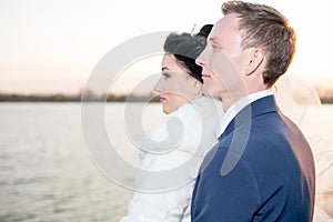 Romantic landscape, the newlywed couple posing at sunset near the river, the bridegroom holds the hand of a bride