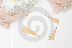 Romantic love letter concept. Flat lay kraft paper envelope, blank note, peony flowers and beige cloth on white wooden table. Top