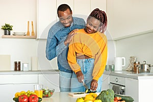 Romantic lunch. Loving black man cuddling wife while woman preparing salad in kitchen, cooking healthy meal together