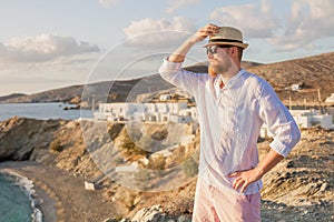 A romantic manly guy with a beard, wearing sunglasses and a hat stands sideways on a rocky shore and looks out to sea