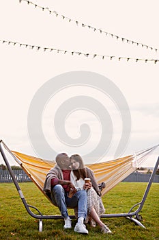 Romantic multiethnic couple with coffee in cup sitting comfortably in hammock in park, covered with cozy plaid, hugging