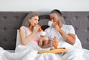 Romantic multiracial couple eating fresh croissants wth coffee, having breakfast in bed at home