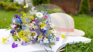 Romantic old-fashioned composition of a cute bouquet of wild and meadow flowers lying on a book and a sun hat of a young girl. A