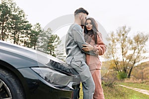 Romantic Ñouple dressed in tracksuits hugs while standing near car in nature.