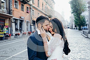 Romantic outdoor portrait of the charming newlyweds hugging. The beautiful brunette bride is tenderly stroking the face