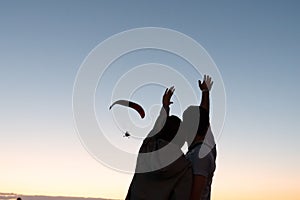 Romantic photo, young couple standing together and waving hands to the hand glider that flying in the sky at sunset