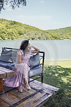 Romantic portrait of beautiful woman in sunglasses and long pink polka dot dress sitting on bench with pillows and looking at