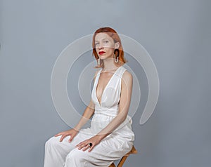 Romantic red-haired woman in white sitting on a wooden chair. Massive jewelry on the ears. Red lipstick