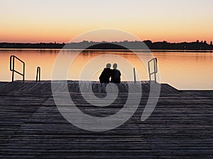 Romantic scene, Young couple sitting on the pier by the lake during sunset. A man and a woman, embracing, look at the