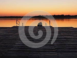 Romantic scene, Young couple sitting on the pier by the lake during sunset. A man and a woman, embracing, look at the