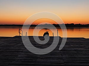 Romantic scene, Young couple sitting on the pier by the lake during sunset. A man and a woman, embracing, look at the