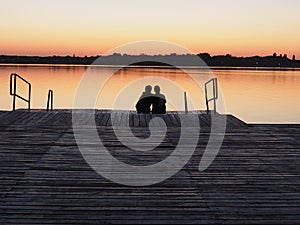 Romantic scene, Young couple sitting on the pier by the lake during sunset. A man and a woman, embracing, look at the