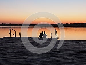 Romantic scene, Young couple sitting on the pier by the lake during sunset. A man and a woman, embracing, look at the
