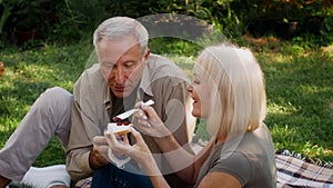 Romantic Senior Couple Having Picnic Outdoors, Sitting On Blanket And Eating Toasts