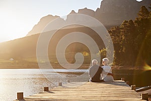 Romantic Senior Couple Sitting On Wooden Jetty By Lake