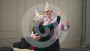 Romantic senior mature couple in party hat cap celebrating birthday anniversary dancing to music together at home. Happy