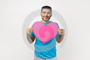 Romantic smiling joyful man standing holding big pink heart, expressing fondness and love.