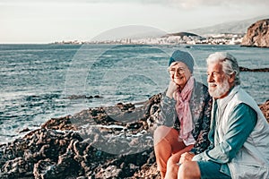 Romantic smiling senior couple sitting on the rocky beach at sea enjoying vacation and retirement at sunset light. Relaxed couple