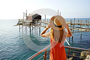 Romantic sunset in Italy. Back view of young woman with hat and dress enjoying view of Trabucco an old fishing machine on Adriatic