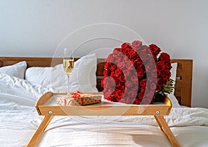 Romantic surprise on Valentine Day. Wrapped gift, bouquet of red roses and glass of champagne on tray on bed.