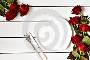 Romantic table setting. Empty heart-shaped plate and cutlery with bouquet of roses on white wooden background. Menu. Flat lay.