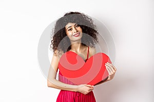 Romantic tender woman with curly hair, hugging big red heart and smiling, look with love, standing against white