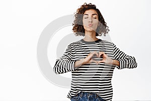 Romantic valentines day. Beautiful young curly girl showing heart hand gesture, pucker lips kissing, standing in casual