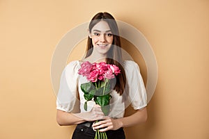 Romantic Valentines day concept. Beautiful young lady holding pink roses and smiling, standing happy on beige background