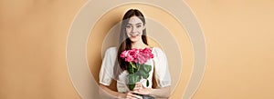 Romantic Valentines day concept. Beautiful young lady holding pink roses and smiling, standing happy on beige background