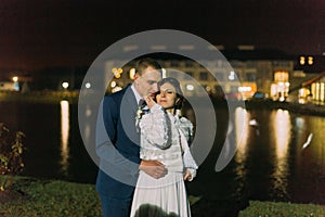 Romantic wedding. Bride and groom posing near night pond illuminated with bright light from banquet hall windows