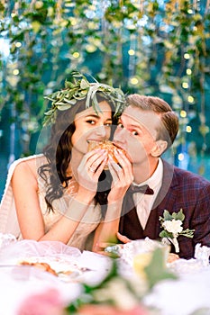 Romantic wedding couple eating a burger together at a rustic restaurant outdoors in forest, looking at camera. Couple