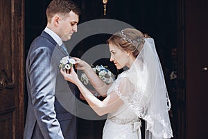 Romantic wedding moment, beautiful blonde bride putting on boutonniere on handsome groom near church entrance, newlywed couple