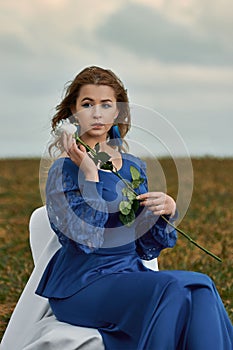 Romantic woman portrait. female in blue dress holds white rose in her hands while sitting on chair in the field