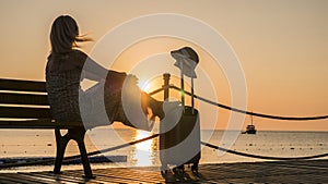 Romantic woman sitting by the sea with a travel bag at sunset. A ship is visible in the distance