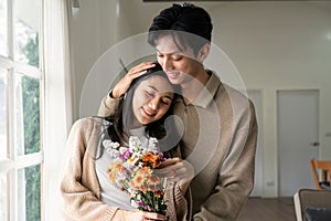 Romantic young asian couple embracing with holding flowers and smiling in living room at home. fall in love. Valentine