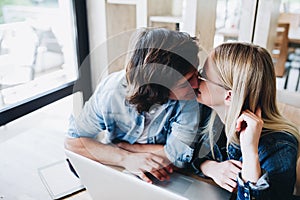 Romantic young couple kissing while sitting at cafe and using la